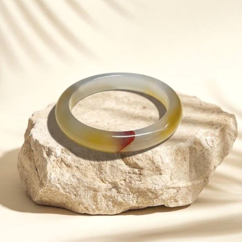 May include: A translucent jade bangle bracelet with a gradient of colors, including yellow, gray, and a touch of red. The bracelet is resting on a textured, light-colored stone. This piece is a beautiful example of natural gemstone jewelry.