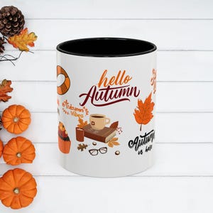May include: White ceramic mug with a black interior, decorated with autumn-themed graphics. The mug features the words "hello Autumn" in a script font, along with images of a coffee cup, leaves, and a cupcake. The mug is surrounded by small pumpkins and fall foliage.