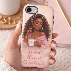 May include: A phone case featuring an illustration of a woman with curly hair, wearing a pink top and gold jewelry, holding a mug. The mug says "Queen". The text "Rested. Blessed. Unbothered." is below the illustration. The case is held by a person.
