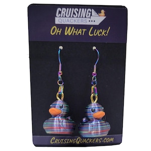 May include: A pair of iridescent rainbow rubber ducky earrings with orange beaks and black eyes. The earrings are hanging from a black card with the text "Cruising Quackers... Oh What Luck!" and "CruisingQuackers.com".