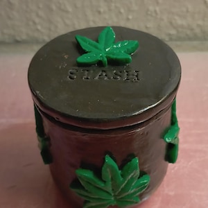 Könnte beinhalten: Ein kleiner, dunkelbrauner Keramiktopf mit Deckel. Der Deckel ist mit einem grünen Cannabisblatt und dem Wort "STASH" verziert. Zusätzliche grüne Blätter verzieren die Seiten des Topfes. Der Topf ist rund und hat eine matte Oberfläche.