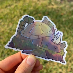 May include: A whimsical sticker featuring a turtle-like creature with a crown, shell, and mossy body. The design incorporates shades of green, purple, and brown, with a touch of iridescent color. The sticker is held against a blurred green background.