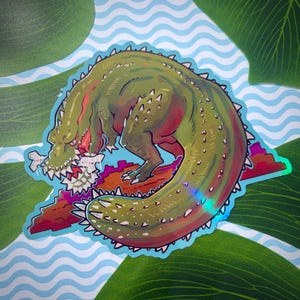 May include: A die-cut sticker featuring a stylized green crocodile with sharp white teeth, biting into a white bone. The creature has a textured, scaly back with spikes, positioned over a reddish-brown horizontal shape with a holographic finish.