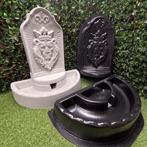 May include: Two wall-mounted fountains featuring a sculpted lion head wearing a crown. One fountain is light gray with a textured finish, and the other is black with a smooth, glossy finish. Each fountain has a semi-circular basin at the base.