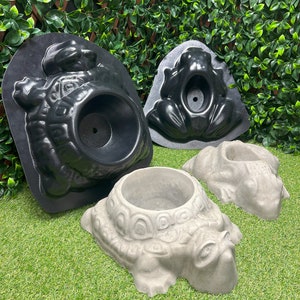 May include: A selection of turtle and frog-shaped planters in different stages of production. Two black moulds are shown, alongside two finished grey concrete planters. These planters are designed for outdoor use and are suitable for small plants.