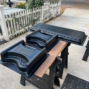 Peut inclure: Moules en plastique noir pour créer des marches décoratives en béton et une bordure rectangulaire. Les moules présentent des volutes ornées et une surface texturée. Les articles sont exposés sur une planche de bois soutenue par des tréteaux noirs.