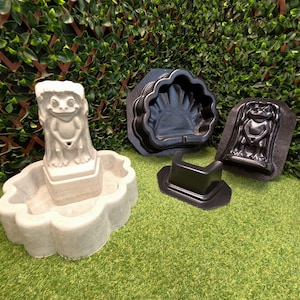 May include: A light gray concrete frog fountain with a scalloped basin, accompanied by three black plastic molds. The molds include a shell-shaped piece, a rectangular base piece, and a detailed frog figure mold for casting concrete garden decor.