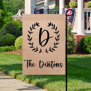 May include: A burlap garden flag featuring a black wreath with the letter "D" in the centre. The text "The Dentons" is printed below the wreath.