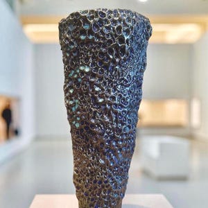 Puede incluir: Un jarrón de cerámica azul oscuro con una superficie texturizada con forma de burbujas. El jarrón es alto y estrecho, con una base redondeada.
