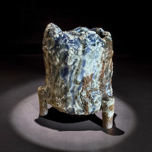 Può includere: Un vaso in ceramica strutturato con una superficie ruvida e irregolare. I colori includono sfumature di blu, marrone e bianco. Il vaso è rialzato su tre brevi gambe cilindriche. La forma generale è cilindrica con un bordo irregolare.