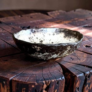 Può includere: Una ciotola in ceramica fatta a mano con un esterno marrone scuro strutturato e un interno bianco sporco macchiettato. La ciotola ha un bordo organico e irregolare e poggia su una superficie di legno scuro.
