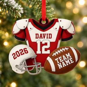 May include: A red and white football jersey ornament with the name DAVID and number 12, accompanied by a white helmet with the year 2026 and a brown football with the text TEAM NAME, all suspended by a red ribbon for holiday decoration.