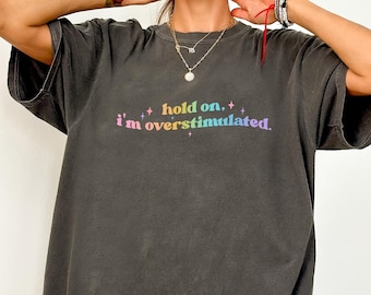 Puede incluir: Una camiseta gris oscura de gran tamaño con el texto "hold on. i'm overstimulated." impreso en colores arcoíris.