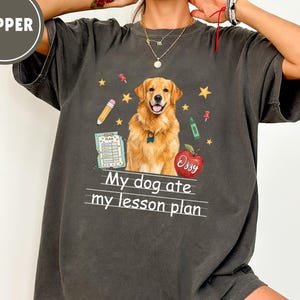 Puede incluir: Una camiseta gris carb&oacute;n de manga corta con un gr&aacute;fico de un perro golden retriever, una manzana roja con el nombre Ozzy, un l&aacute;piz y un documento de plan de lecci&oacute;n. El texto My dog ate my lesson plan est&aacute; impreso en blanco bajo el perro.