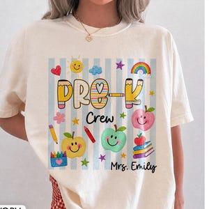 May include: An ivory short-sleeved t-shirt featuring a graphic print with light blue vertical stripes. The design includes the text "PRE-K Crew" and "Mrs. Emily" surrounded by colorful illustrations of smiling apples, pencils, stars, and a rainbow.