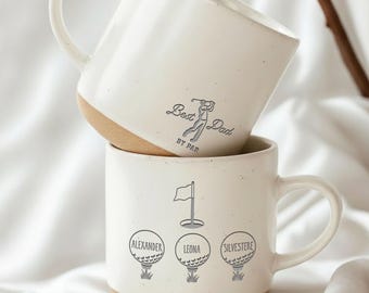 Puede incluir: Dos tazas de cerámica blanca con base beige apiladas. La taza superior tiene impreso "Best Dad by Par", con un gráfico de golfista. La taza inferior tiene una bandera de golf y tres diseños de pelotas de golf con nombres.