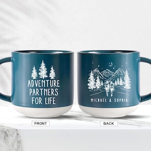 May include: Two teal blue ceramic mugs with white text and illustrations. The mug on the left says "Adventure Partners For Life". The mug on the right has a mountain scene with two figures hiking and says "Michael & Sophia".