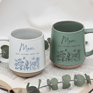 May include: Two ceramic mugs, one white and one green, with the word "Mom" in script. The white mug has blue floral illustrations and the names "Jack - Victoria - Rose - Kai". The green mug has similar floral illustrations.