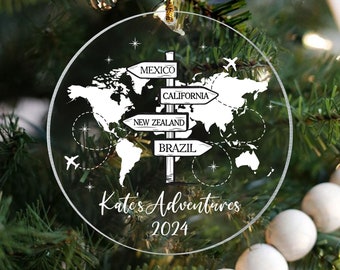 Puede incluir: Adorno navideño redondo y transparente con un diseño de mapa del mundo y señales direccionales que apuntan a México, California, Nueva Zelanda y Brasil. También aparece el texto "Kate's Adventures 2024". El adorno cuelga de un hilo dorado.