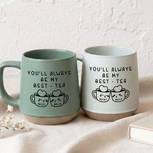 May include: Two ceramic mugs, one in mint green and one in light blue, with a beige base. Each mug features a black graphic of two smiling teacups with hearts and the text "YOU'LL ALWAYS BE MY BEST-TEA". The names "BECKY" and "BELLA" are below the teacups.
