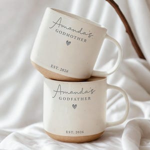 May include: Two stacked white ceramic mugs with a speckled finish and tan bottoms. The top mug reads "Amanda's Godmother" with a gray heart and "EST. 2026." The bottom mug says "Amanda's Godfather" with a gray heart and "EST. 2026."