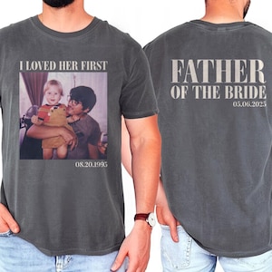 May include: A gray t-shirt with the text "I loved her first" and a photo of a young girl and a man. The date "08.20.1995" is printed below the photo. The back of the shirt has the text "Father of the Bride" and the date "05.06.2025".