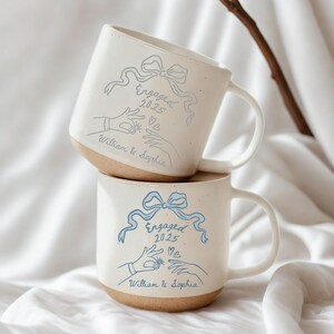 May include: Two stacked white ceramic mugs with a light brown base. Each mug has a blue ribbon design with the words "Engaged 2025", hands, and the names "William & Sophia". The mugs are on a white background.