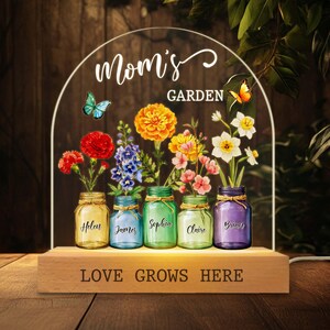 May include: A decorative LED light featuring a wooden base with the text "LOVE GROWS HERE." Above, a clear arch displays the words "Mom's GARDEN" and colorful flowers in mason jars. Butterflies and names are also visible.