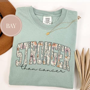 May include: A light green t-shirt with the words "STRONGER than cancer" in a floral design. The shirt has a crew neck and short sleeves. A gold necklace with a small pendant is draped across the shirt.