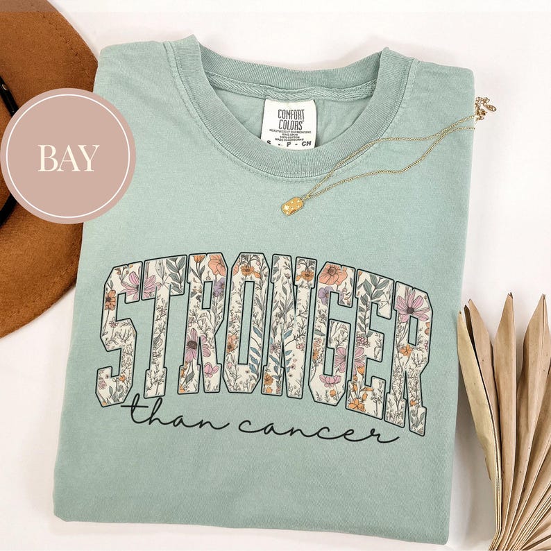 May include: A light green t-shirt with the words "STRONGER than cancer" in a floral design. The shirt has a crew neck and short sleeves. A gold necklace with a small pendant is draped across the shirt.