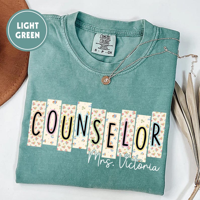 May include: A light green Comfort Colors t-shirt with the word "COUNSELOR" in black letters with floral accents. The name "Mrs. Victoria" is printed below. A silver necklace with a pendant is draped on the shirt.