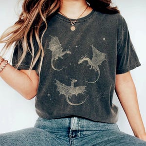 K&ouml;nnte beinhalten: Dunkelgraues T-Shirt mit einem Design von drei Drachen mit ausgebreiteten Fl&uuml;geln und Sternen. Die Drachen sind hellbraun. Das Shirt hat kurze &Auml;rmel und einen Rundhalsausschnitt.