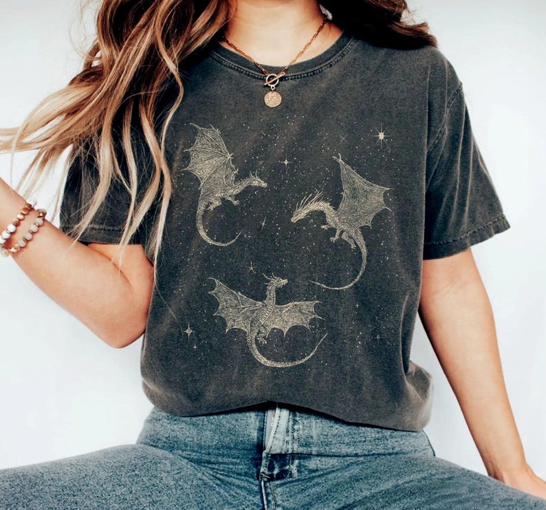 K&ouml;nnte beinhalten: Dunkelgraues T-Shirt mit einem Design von drei Drachen mit ausgebreiteten Fl&uuml;geln und Sternen. Die Drachen sind hellbraun. Das Shirt hat kurze &Auml;rmel und einen Rundhalsausschnitt.