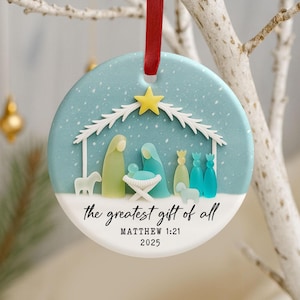 May include: Round Christmas ornament with a light blue background and a red ribbon. The ornament features a nativity scene with a yellow star, figures, and the text "the greatest gift of all" and "MATTHEW 1:21 2025".