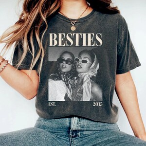 May include: Dark gray t-shirt with the word "BESTIES" in a bold, vintage font. A black and white photo of two women wearing sunglasses and headscarves is printed below the word. The text "EST. 2015" is printed below the photo.