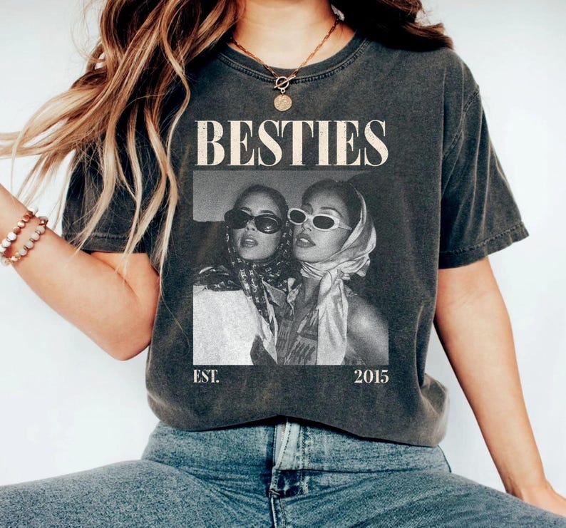 May include: Dark gray t-shirt with the word "BESTIES" in a bold, vintage font. A black and white photo of two women wearing sunglasses and headscarves is printed below the word. The text "EST. 2015" is printed below the photo.