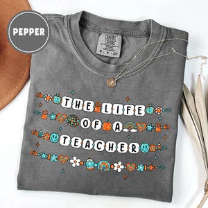 May include: A gray Comfort Colors t-shirt with the text "THE LIFE OF A TEACHER" in white block letters. The shirt features colorful graphics of apples, rainbows, and other school-themed designs. A silver necklace with a pendant is draped on the shirt.
