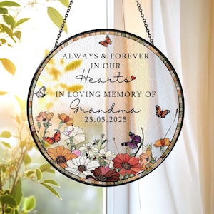 May include: Round stained glass memorial with a black metal frame and chain. The glass features the text "ALWAYS & FOREVER IN OUR Hearts IN LOVING MEMORY OF Grandma 25.05.2025". Colorful flowers and butterflies decorate the border.