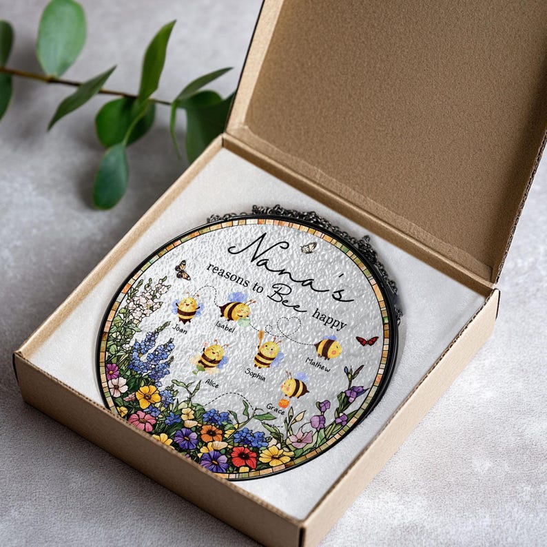 May include: A round, decorative glass piece in a gift box. The glass features a floral and bee design with the text "Nana's reasons to Bee happy" and names. The piece is framed in black and comes in a brown cardboard box.
