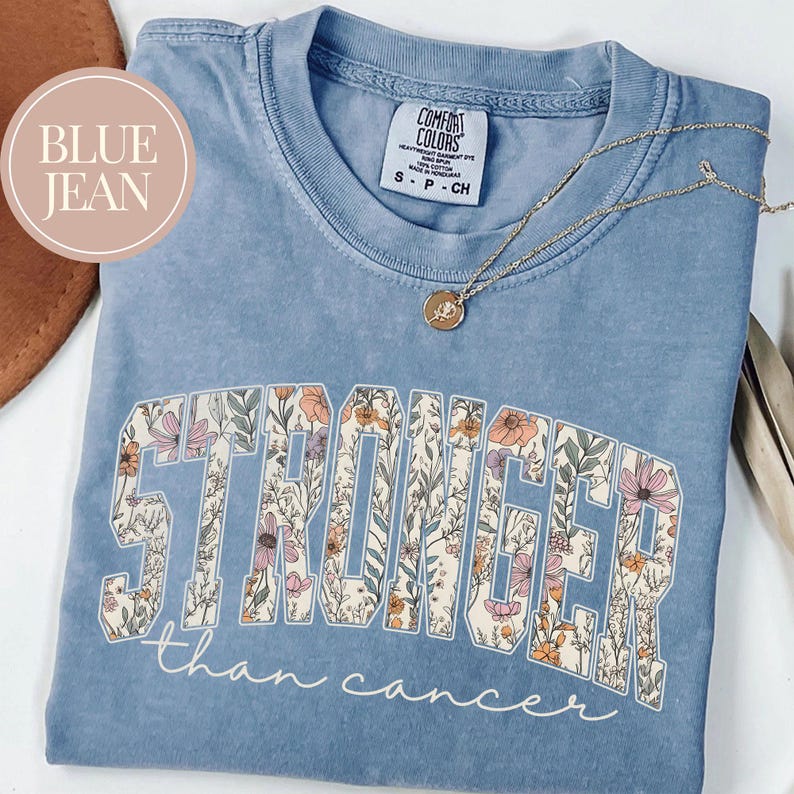May include: A blue jean Comfort Colors t-shirt with the words "STRONGER than cancer" in a floral design. A gold necklace with a pendant is draped across the shirt.
