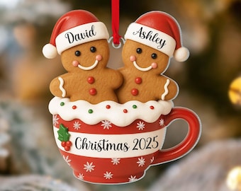 May include: A Christmas ornament featuring two gingerbread people wearing Santa hats, labeled "David" and "Ashley." They sit in a red and white cup with "Christmas 2025" printed on it. A red ribbon is attached for hanging.