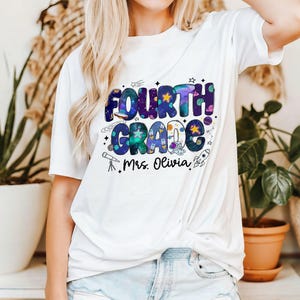 May include: White t-shirt with a colorful "FOURTH GRADE" graphic in a space theme, including stars, planets, and a rocket. The text "Mrs. Olivia" is printed below the main design.