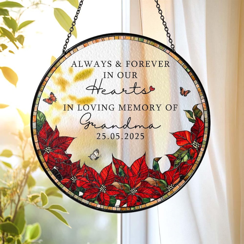 May include: Round stained glass memorial with a black metal frame and chain. The glass features the text "ALWAYS & FOREVER IN OUR Hearts IN LOVING MEMORY OF Grandma 25.05.2025". Red poinsettias and butterflies decorate the edges.