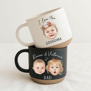 May include: Two ceramic mugs, one white and one black, stacked. The white mug has "I Love You Grandma" with a baby illustration. The black mug has "Ronan & Victoria's Dad" with two baby illustrations. Both mugs have a brown base.