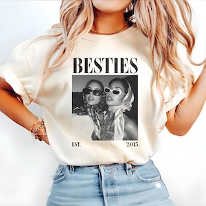 May include: Cream-colored t-shirt featuring the word "BESTIES" in a bold, black font above a black and white photo of two women wearing sunglasses and headscarves. The text "EST. 2015" is printed below the photo.