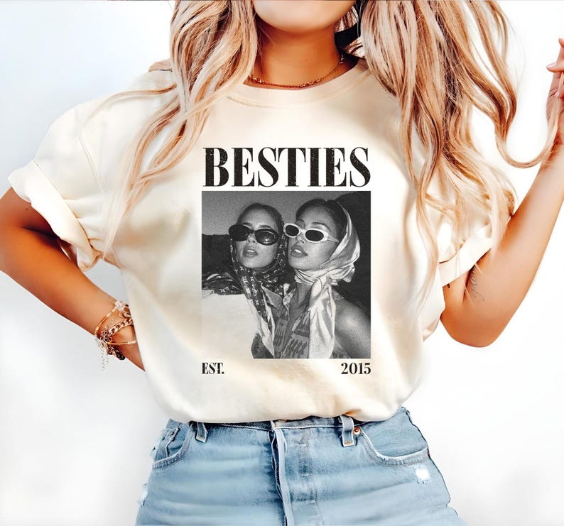 May include: Cream-colored t-shirt featuring the word "BESTIES" in a bold, black font above a black and white photo of two women wearing sunglasses and headscarves. The text "EST. 2015" is printed below the photo.