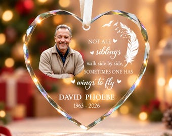 May include: A heart-shaped crystal ornament featuring a photo of a smiling person, a white feather graphic, and the text: NOT ALL siblings walk side by side, SOMETIMES ONE HAS wings to fly, DAVID PHOEBE, 1963 - 2026. It hangs from a white ribbon.