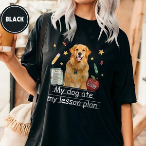 Puede incluir: Una camiseta negra con un gr&aacute;fico de un perro golden retriever rodeado de ilustraciones escolares que incluyen un l&aacute;piz, una manzana roja con el nombre Ozzy, un portapapeles y estrellas amarillas, con el texto My dog ate my lesson plan.