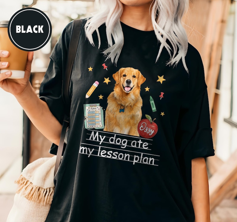 Puede incluir: Una camiseta negra con un gr&aacute;fico de un perro golden retriever rodeado de ilustraciones escolares que incluyen un l&aacute;piz, una manzana roja con el nombre Ozzy, un portapapeles y estrellas amarillas, con el texto My dog ate my lesson plan.