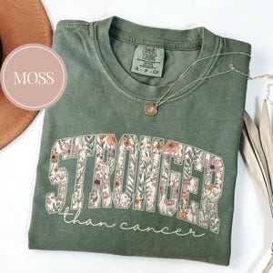 May include: A moss green t-shirt with the word "STRONGER" in large floral letters and the phrase "than cancer" in a script font. A silver necklace with a pendant rests on the shirt. The shirt is folded.